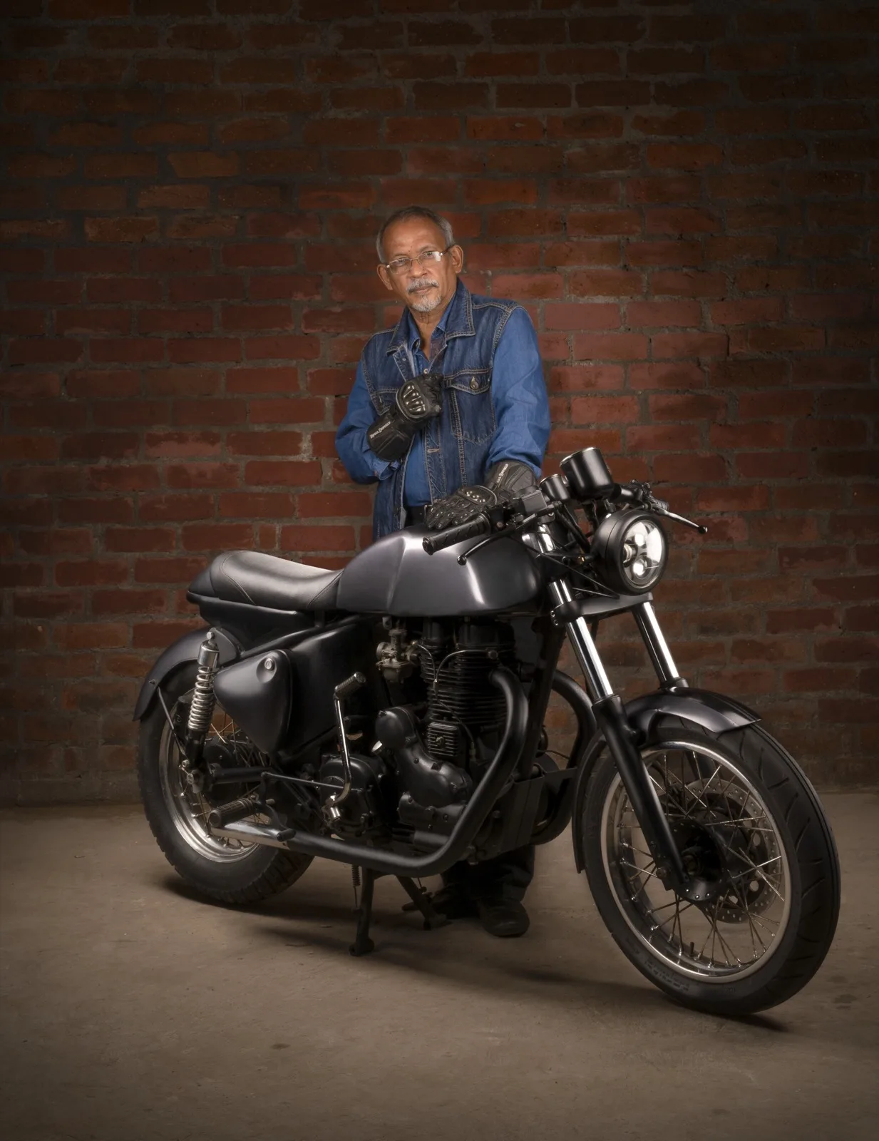 Studio portrait of a man in a denim jacket and riding gloves standing behind a matte-black cafe racer motorcycle against a brick wall